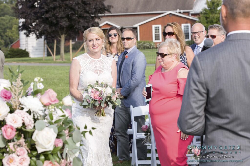 JenniferRudiWeddingCeremonyPhotos-97 - TOMMY GATZ ENTERTAINMENT A bride in a white dress walks down an outdoor aisle, holding a bouquet, as guests watch. A barn and greenery are in the background.