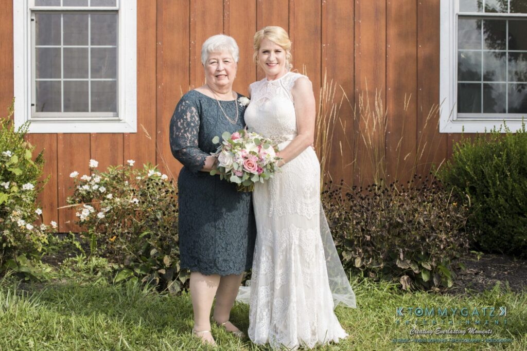 JenniferRudiWeddingFormalPhotos-193 - TOMMY GATZ ENTERTAINMENT Two women stand side by side in front of a wooden building, both smiling. One woman holds a bouquet of flowers.