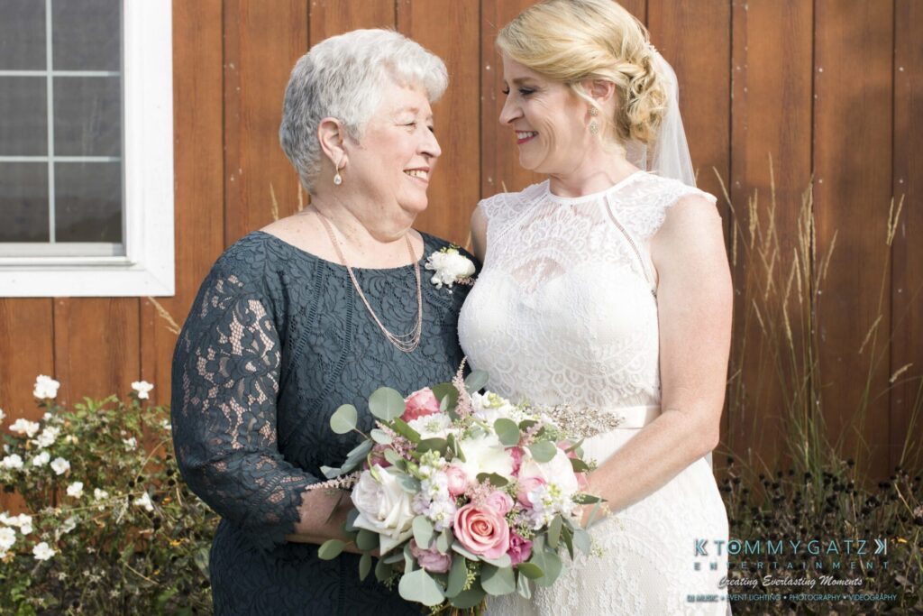 JenniferRudiWeddingFormalPhotos-194 - TOMMY GATZ ENTERTAINMENT Two women facing each other and smiling. One is in a white dress holding a bouquet of flowers, and the other is in a dark embroidered dress with a white corsage. Brown wooden wall in the background.