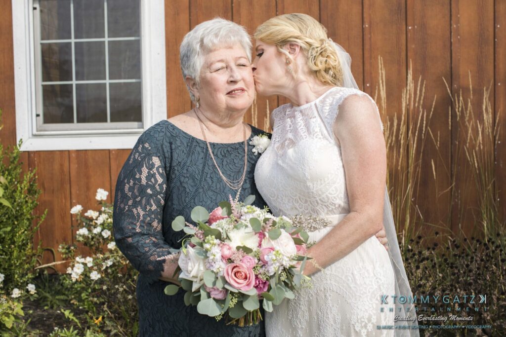 JenniferRudiWeddingFormalPhotos-195 - TOMMY GATZ ENTERTAINMENT A woman in a wedding dress kisses an older woman on the cheek. The older woman holds a bouquet of flowers. They stand in front of a wooden wall with a window.