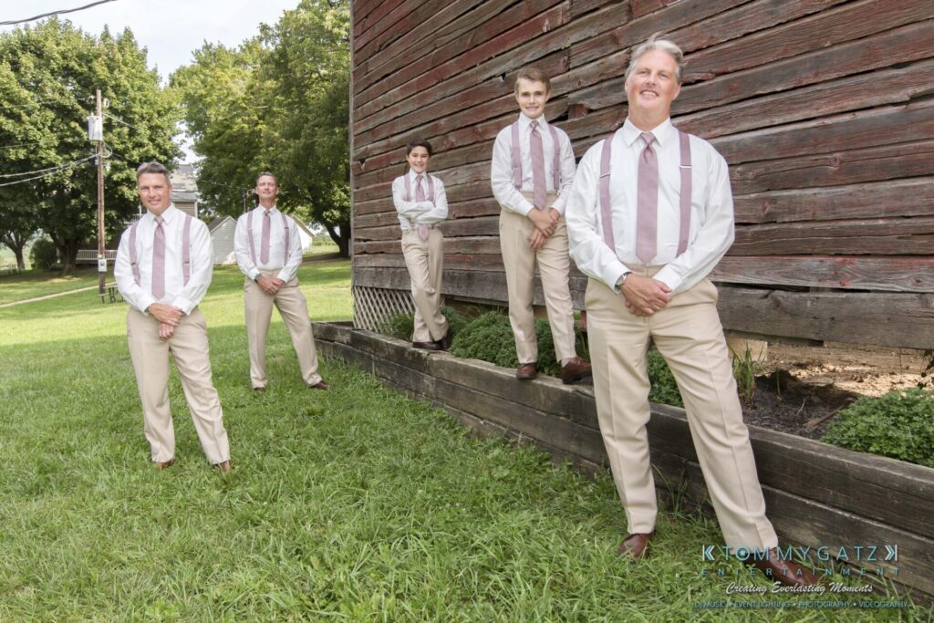 JenniferRudiWeddingFormalPhotos-207 - TOMMY GATZ ENTERTAINMENT Five men in matching beige suits and pink ties pose in front of a wooden building on a grassy area.