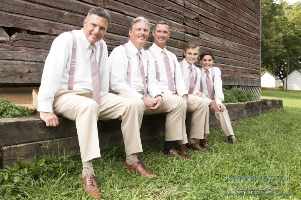 JenniferRudiWeddingFormalPhotos-208 - TOMMY GATZ ENTERTAINMENT Five people in light shirts and khaki pants sit on a wooden beam outside a rustic building, posing for a photo on a grassy area.