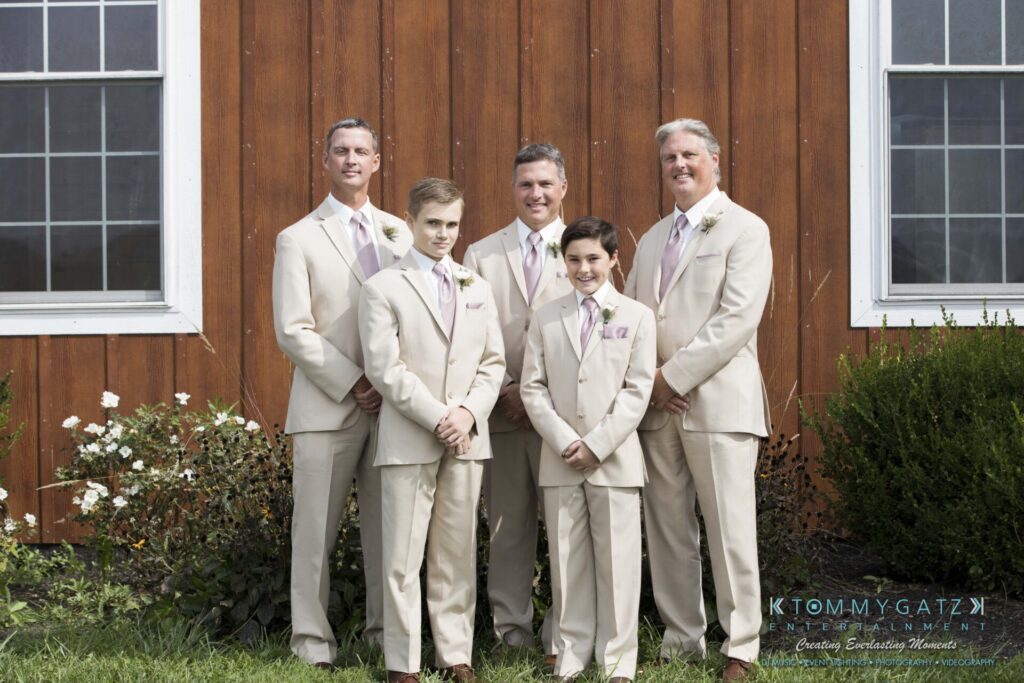 JenniferRudiWeddingPrepPhotos-18 - TOMMY GATZ ENTERTAINMENT Five men in beige suits stand together in front of a wooden building, with green grass and plants around them.