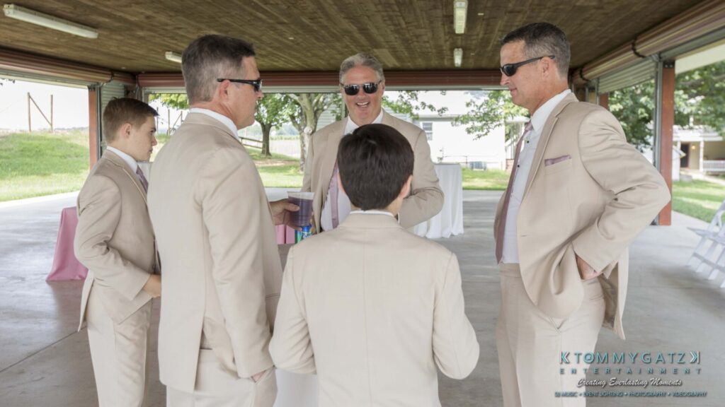 JenniferRudiWeddingPrepPhotos-2 - TOMMY GATZ ENTERTAINMENT Five men in beige suits and sunglasses stand together in a shaded outdoor area, conversing and smiling.
