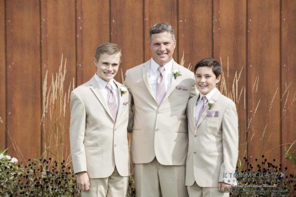 JenniferRudiWeddingPrepPhotos-21 - TOMMY GATZ ENTERTAINMENT Three people in beige suits with lavender ties stand in front of a wooden wall, smiling.