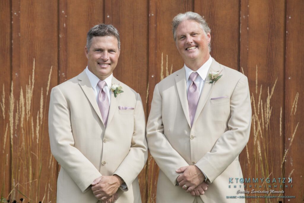 JenniferRudiWeddingPrepPhotos-26 - TOMMY GATZ ENTERTAINMENT Two men in beige suits with pink ties stand side by side in front of a wooden backdrop, smiling at the camera.