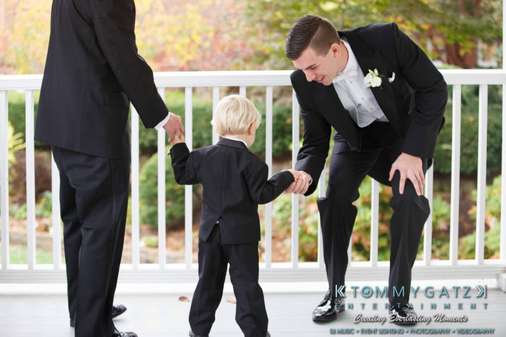 JulieKyleLongWedding11715-167 - TOMMY GATZ ENTERTAINMENT A young boy in a suit fist-bumps an adult man in a tuxedo on a porch, with another adult standing nearby.