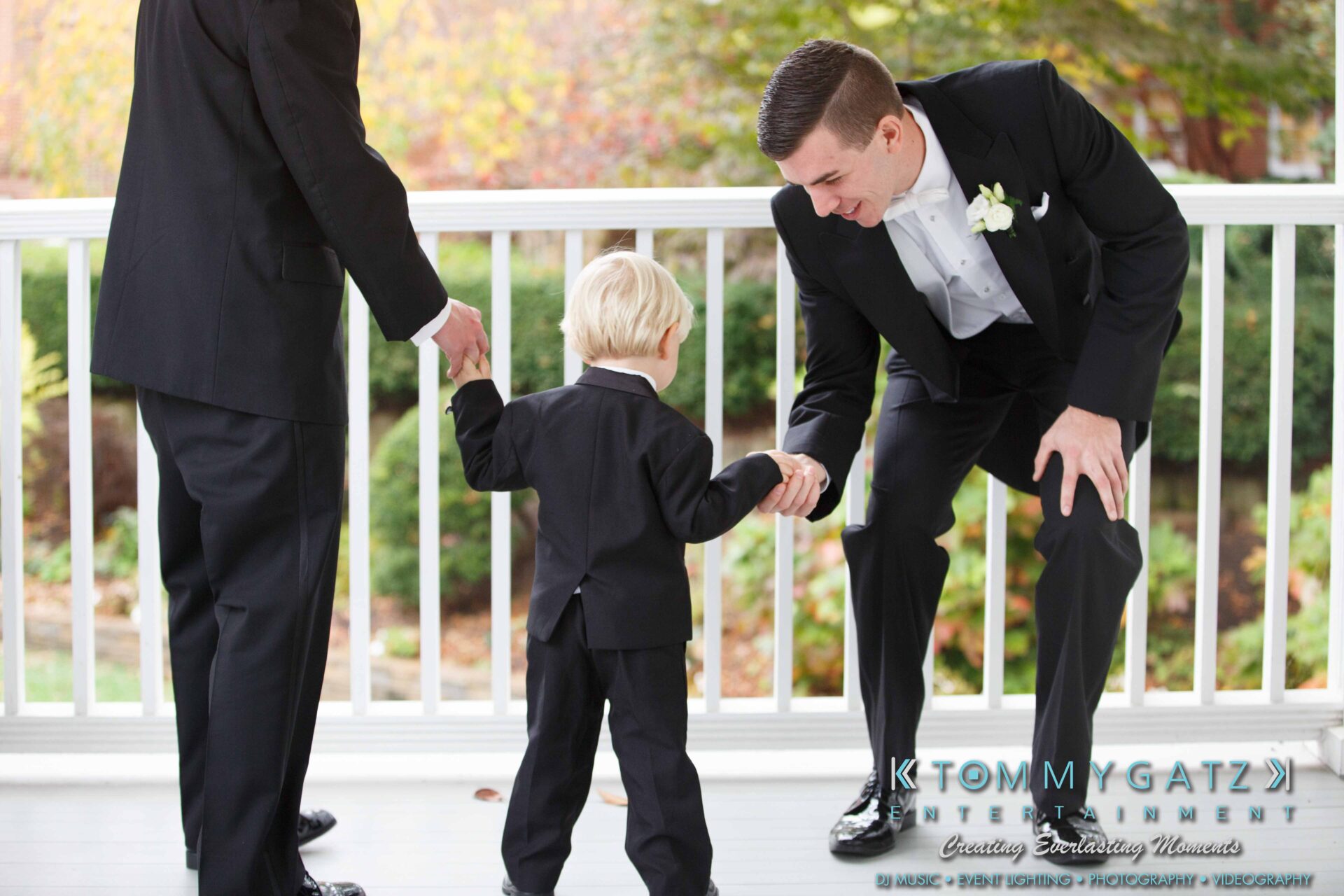 JulieKyleLongWedding11715-167 - TOMMY GATZ ENTERTAINMENT A young boy in a suit fist-bumps an adult man in a tuxedo on a porch, with another adult standing nearby.