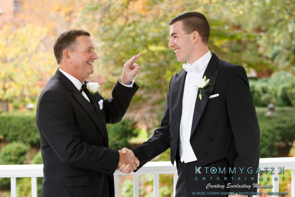 JulieKyleLongWedding11715-178 - TOMMY GATZ ENTERTAINMENT Two men in tuxedos shake hands and smile on a covered porch. One man points at the other. Greenery and fall foliage are visible in the background.