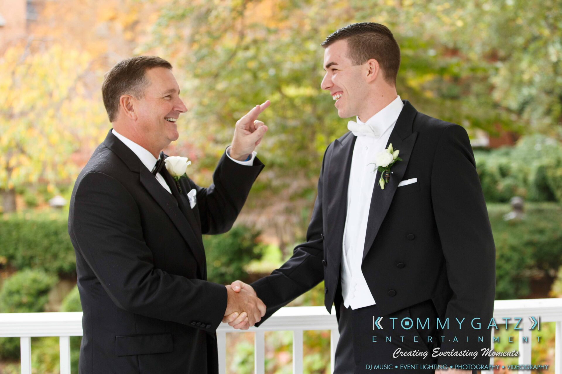 JulieKyleLongWedding11715-178 - TOMMY GATZ ENTERTAINMENT Two men in tuxedos shake hands and smile on a covered porch. One man points at the other. Greenery and fall foliage are visible in the background.