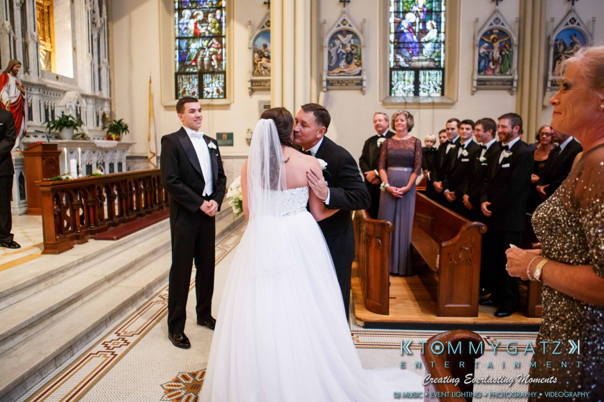 JulieKyleLongWedding11715-220 - TOMMY GATZ ENTERTAINMENT A bride and groom kiss at the altar in a church. A groomsman and guests look on, with stained glass windows in the background.
