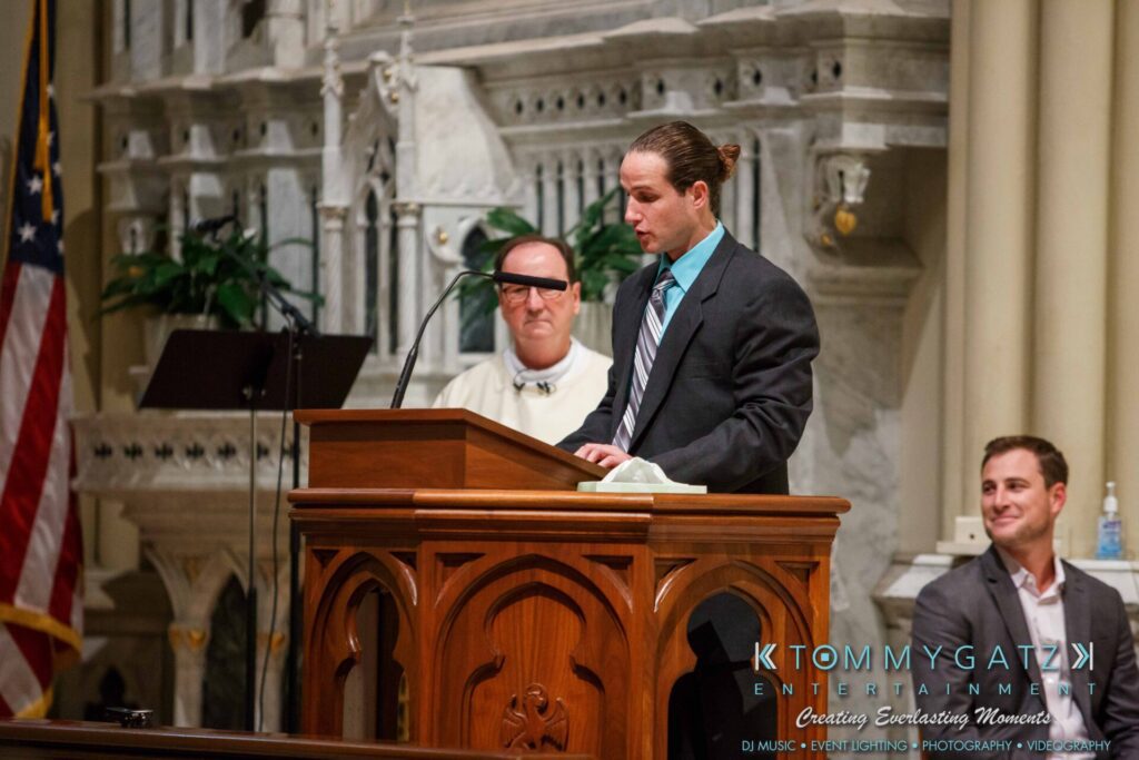 JulieKyleLongWedding11715-237 - TOMMY GATZ ENTERTAINMENT A man in a suit speaks at a church podium. Another man in white robes and a seated man in a suit are in the background. The setting is ornate with stone architecture.