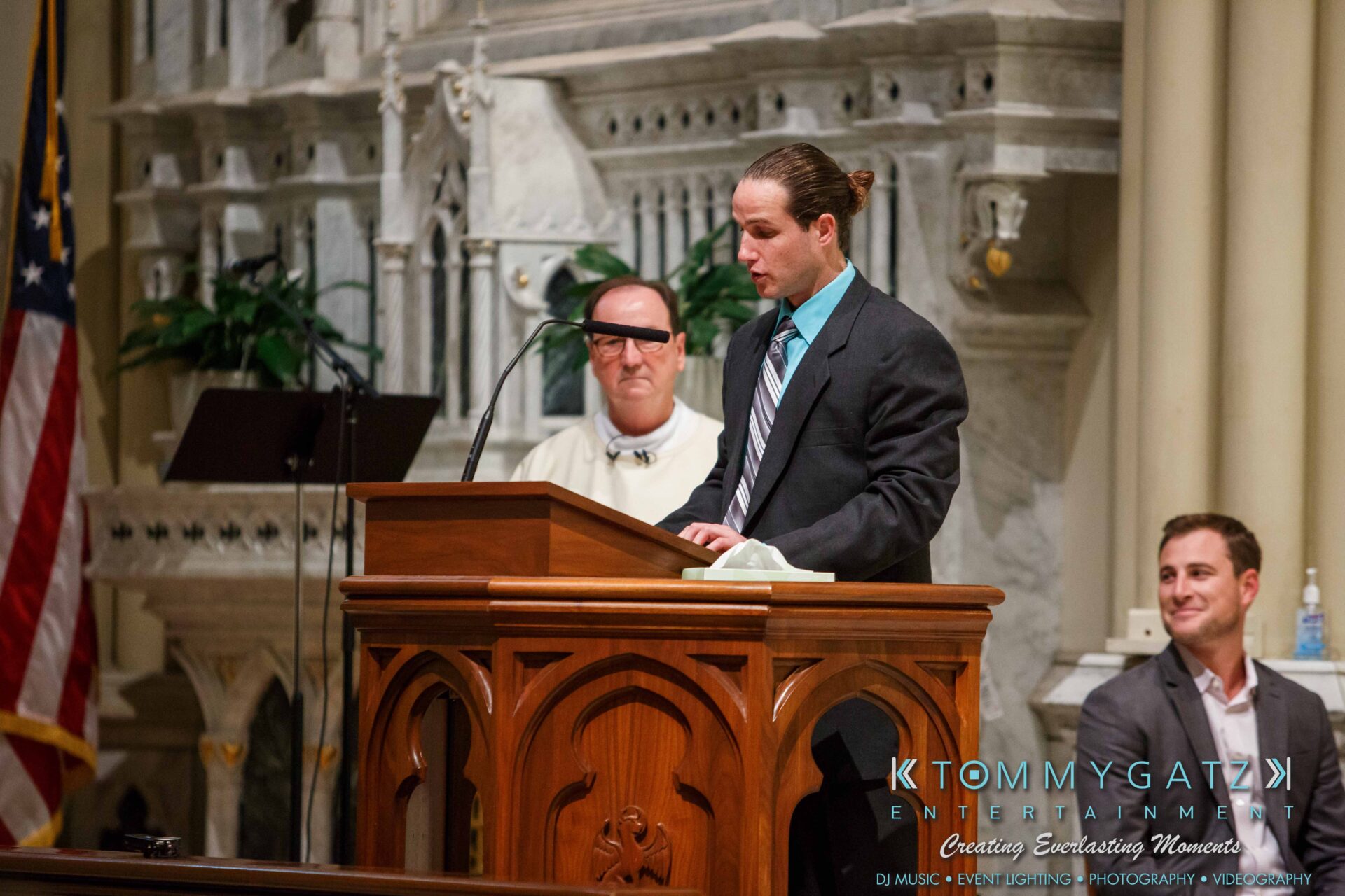 JulieKyleLongWedding11715-237 - TOMMY GATZ ENTERTAINMENT A man in a suit speaks at a church podium. Another man in white robes and a seated man in a suit are in the background. The setting is ornate with stone architecture.