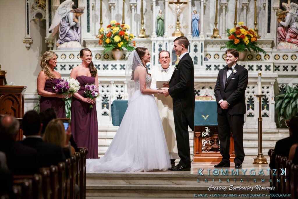 JulieKyleLongWedding11715-261 - TOMMY GATZ ENTERTAINMENT A couple stands holding hands at the altar during a wedding ceremony inside a church, with bridesmaids and a groomsman nearby. A priest officiates in front of a decorated altar.