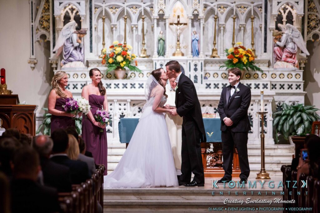 JulieKyleLongWedding11715-271 - TOMMY GATZ ENTERTAINMENT A couple shares a kiss at their wedding ceremony in a church with an altar in the background. Bridesmaids and a groomsman stand nearby.