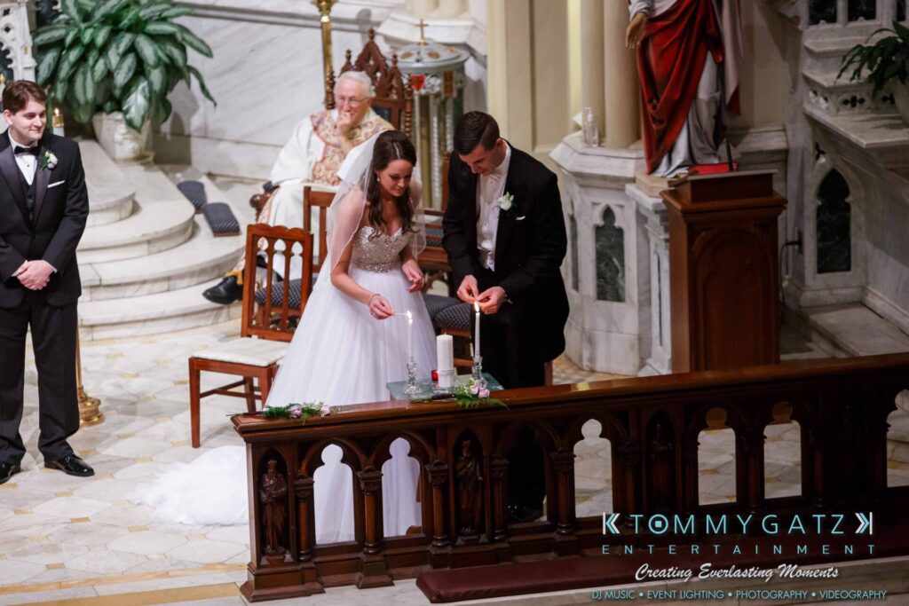 JulieKyleLongWedding11715-278 - TOMMY GATZ ENTERTAINMENT Bride and groom lighting a unity candle during a wedding ceremony in a church, with a priest and groomsmen nearby.