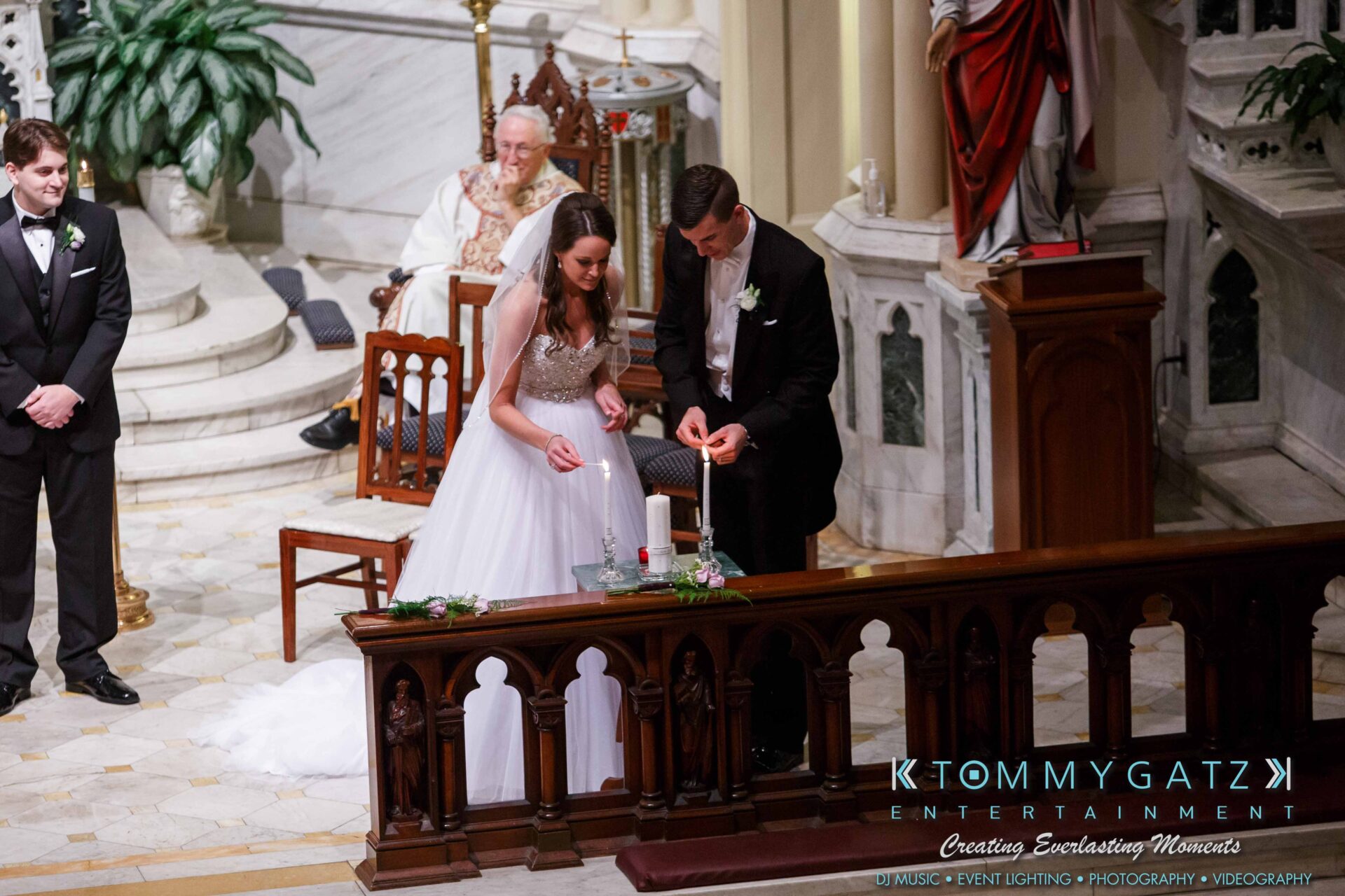 JulieKyleLongWedding11715-278 - TOMMY GATZ ENTERTAINMENT Bride and groom lighting a unity candle during a wedding ceremony in a church, with a priest and groomsmen nearby.