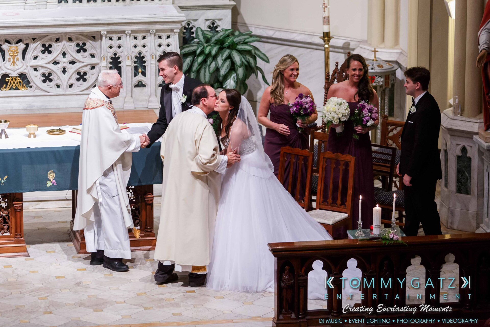 JulieKyleLongWedding11715-299 - TOMMY GATZ ENTERTAINMENT A wedding ceremony in a church with a bride and groom exchanging vows, surrounded by clergy and bridesmaids holding bouquets.