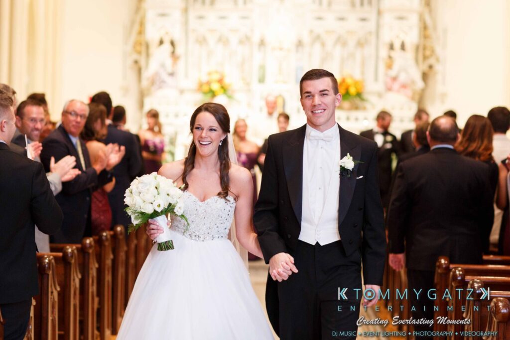 JulieKyleLongWedding11715-335 - TOMMY GATZ ENTERTAINMENT A couple walks down the aisle at a wedding ceremony, smiled at by guests. The bride holds a bouquet and wears a white gown, while the groom wears a tuxedo.