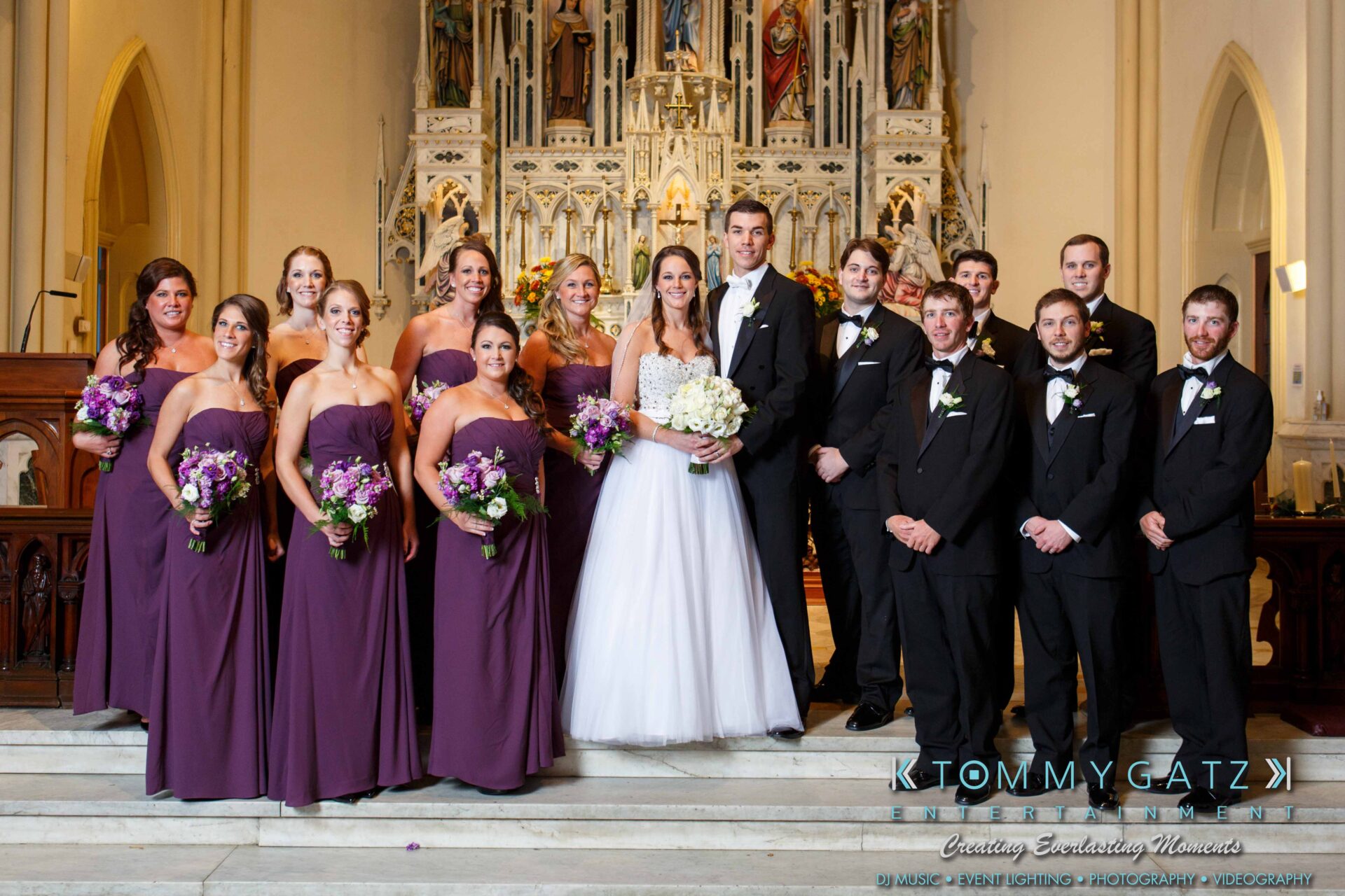 JulieKyleLongWedding11715-350 - TOMMY GATZ ENTERTAINMENT A wedding party poses in a church. The bride and groom stand in the center, surrounded by bridesmaids in purple dresses and groomsmen in black suits.
