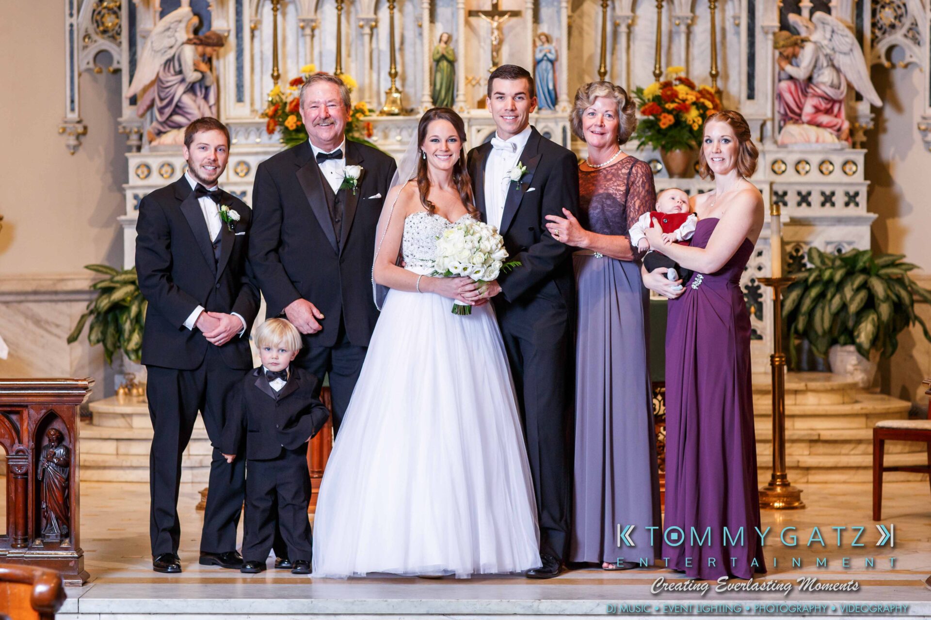 JulieKyleLongWedding11715-358 - TOMMY GATZ ENTERTAINMENT A wedding party stands together in a church, with the bride and groom at the center. A child stands in front, and family members surround them, dressed formally.