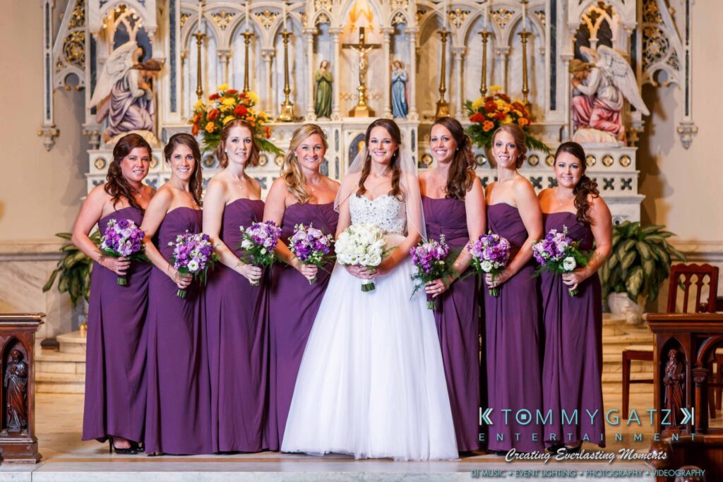JulieKyleLongWedding11715-367 - TOMMY GATZ ENTERTAINMENT Bride in white with bridesmaids in purple dresses holding bouquets, standing in a decorated church.