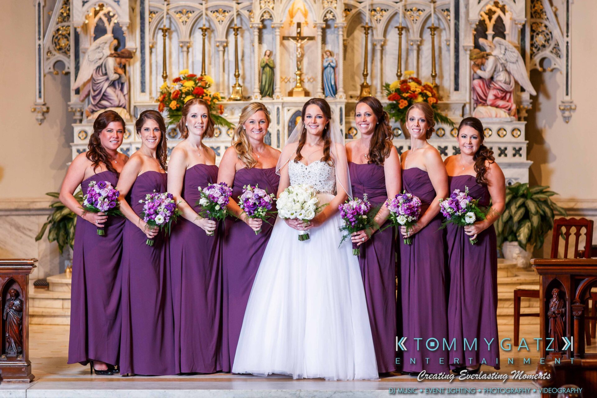 JulieKyleLongWedding11715-367 - TOMMY GATZ ENTERTAINMENT Bride in white with bridesmaids in purple dresses holding bouquets, standing in a decorated church.
