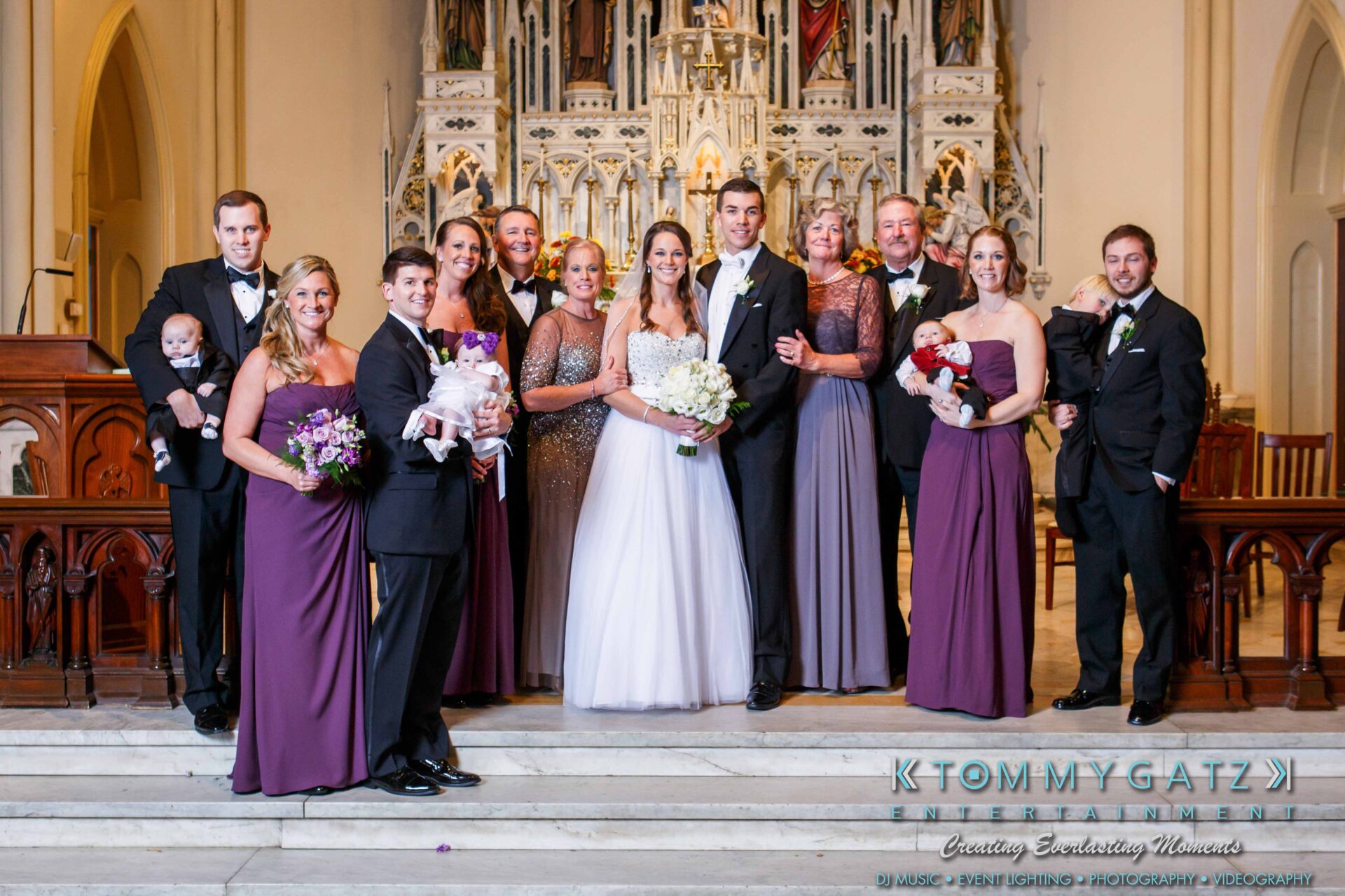 JulieKyleLongWedding11715-371 - TOMMY GATZ ENTERTAINMENT A wedding party poses in front of an ornate altar. The group includes the bride and groom, bridesmaids in purple dresses, groomsmen in suits, and two infants held by women.