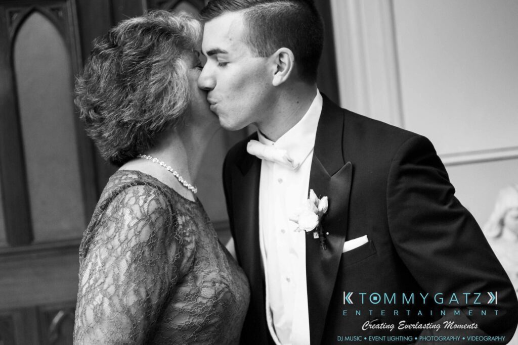 JulieKyleLongWedding11715-46 - TOMMY GATZ ENTERTAINMENT A groom kisses a woman on the cheek, both in formal attire, against a backdrop of wood and decorative paneling.