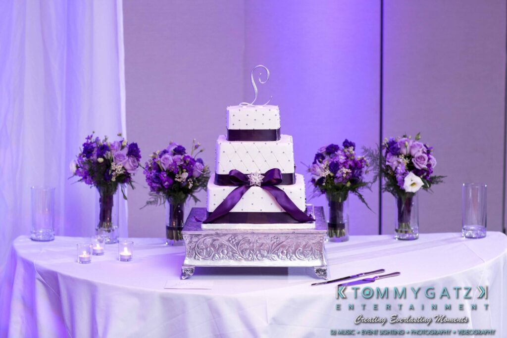 JulieKyleLongWedding11715-609 - TOMMY GATZ ENTERTAINMENT Three-tiered white cake with purple ribbon on a silver stand, surrounded by purple flower arrangements and tea lights on a white tablecloth.