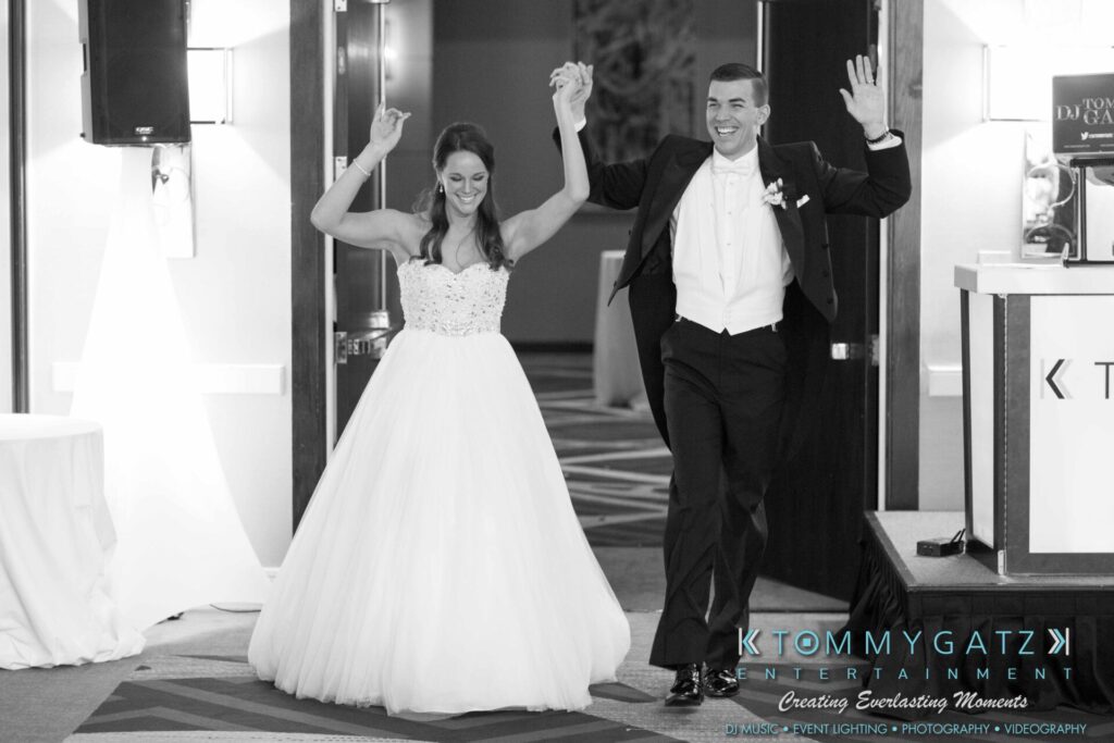 JulieKyleLongWedding11715-635 - TOMMY GATZ ENTERTAINMENT A bride and groom joyfully entering a wedding reception hall, holding hands and smiling.