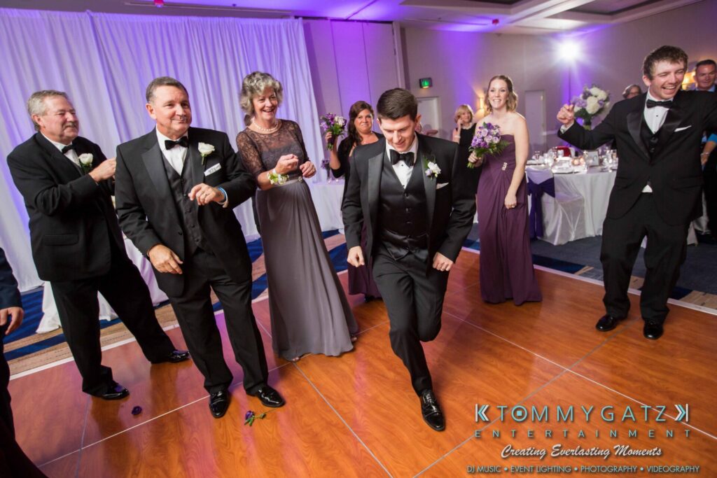 JulieKyleLongWedding11715-647 - TOMMY GATZ ENTERTAINMENT A group of people in formal attire dancing energetically at an indoor event with a wooden floor and purple lighting.