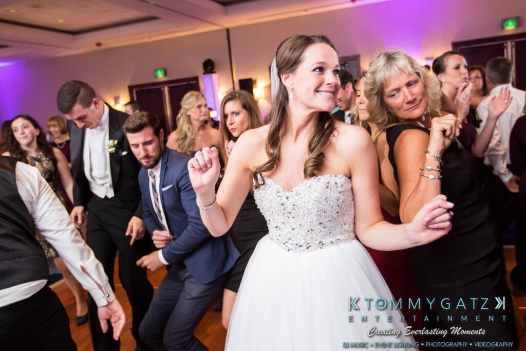 JulieKyleLongWedding11715-671 - TOMMY GATZ ENTERTAINMENT People dancing energetically at a formal event, with a bride in a white dress leading the group.