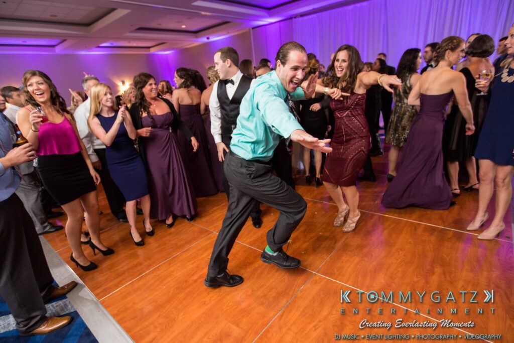 JulieKyleLongWedding11715-685 - TOMMY GATZ ENTERTAINMENT A group of people dancing energetically on a wooden floor at an indoor event, with purple lighting in the background.