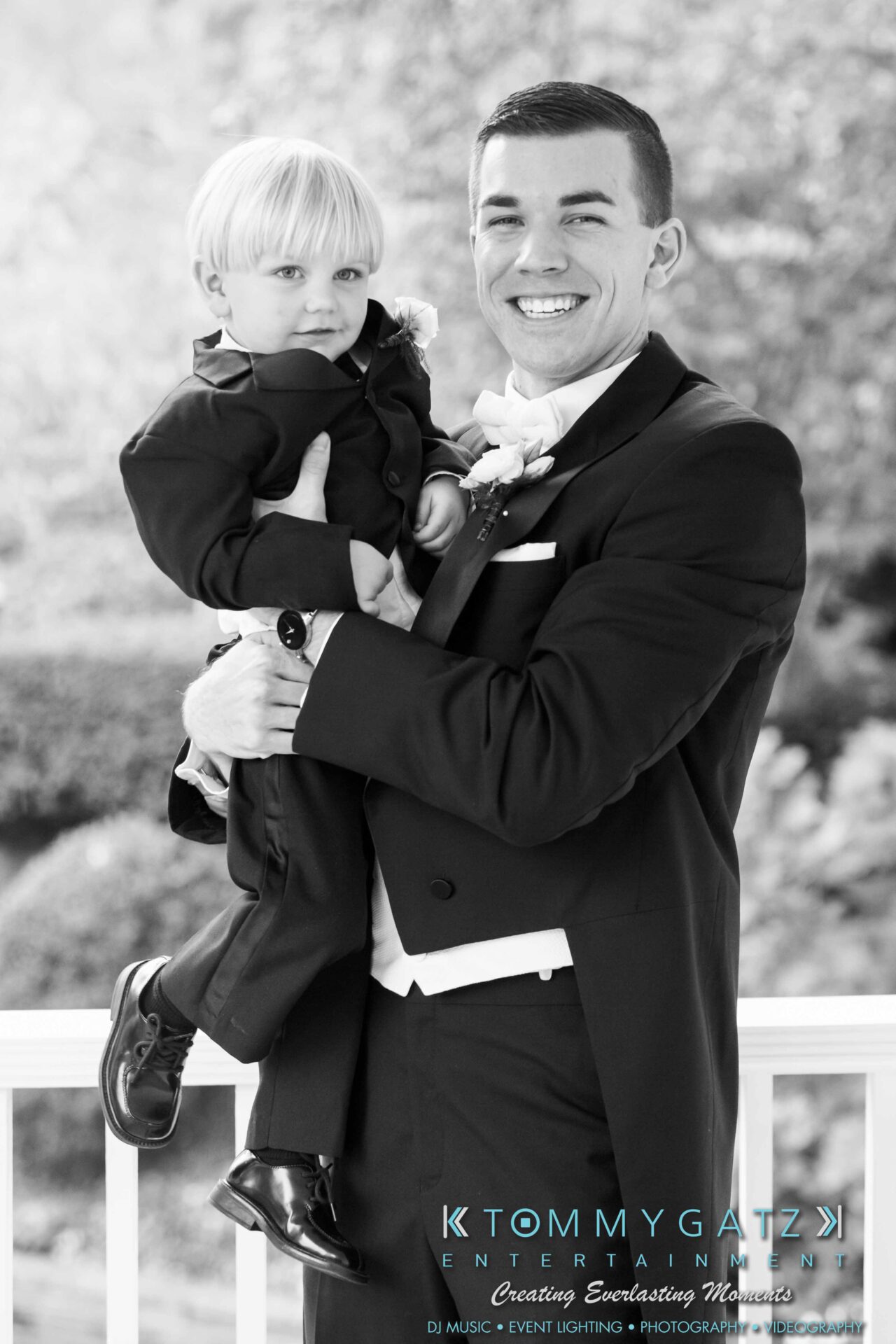 JulieKyleLongWedding11715-76 - TOMMY GATZ ENTERTAINMENT Man in a tuxedo holding a young boy dressed in a suit, both smiling. They're standing on a porch with a blurred garden background.