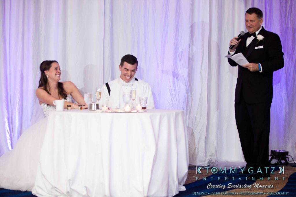 JulieKyleLongWedding11715-760 - TOMMY GATZ ENTERTAINMENT A bride and groom sitting at a table laugh as a man in a tuxedo speaks into a microphone.