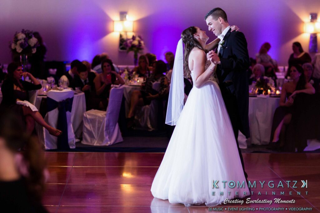 JulieKyleLongWedding11715-786 - TOMMY GATZ ENTERTAINMENT Bride and groom share their first dance in a dimly lit reception hall with guests seated around them.