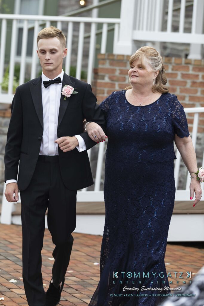Kari-Jose-062318-CEREMONY-Low-Res-WM-207 - TOMMY GATZ ENTERTAINMENT A young man in a suit escorts an older woman in a blue dress. They are walking outdoors on a brick path with a white railing behind them.