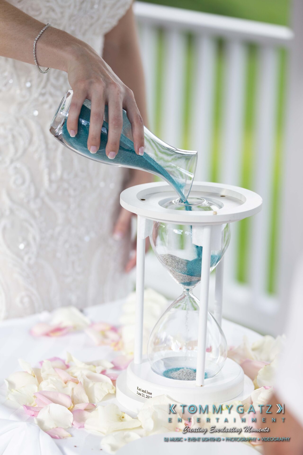 Kari-Jose-062318-CEREMONY-Low-Res-WM-311 - TOMMY GATZ ENTERTAINMENT A person in a white dress pours blue sand into an hourglass on a table with rose petals.