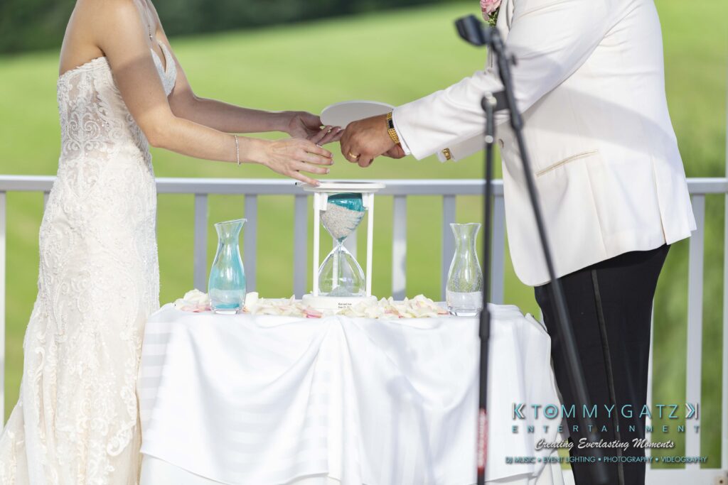 Kari-Jose-062318-CEREMONY-Low-Res-WM-321 - TOMMY GATZ ENTERTAINMENT A bride and groom pour sand into an hourglass during a wedding ceremony, standing behind a table with a white cloth and rose petals.