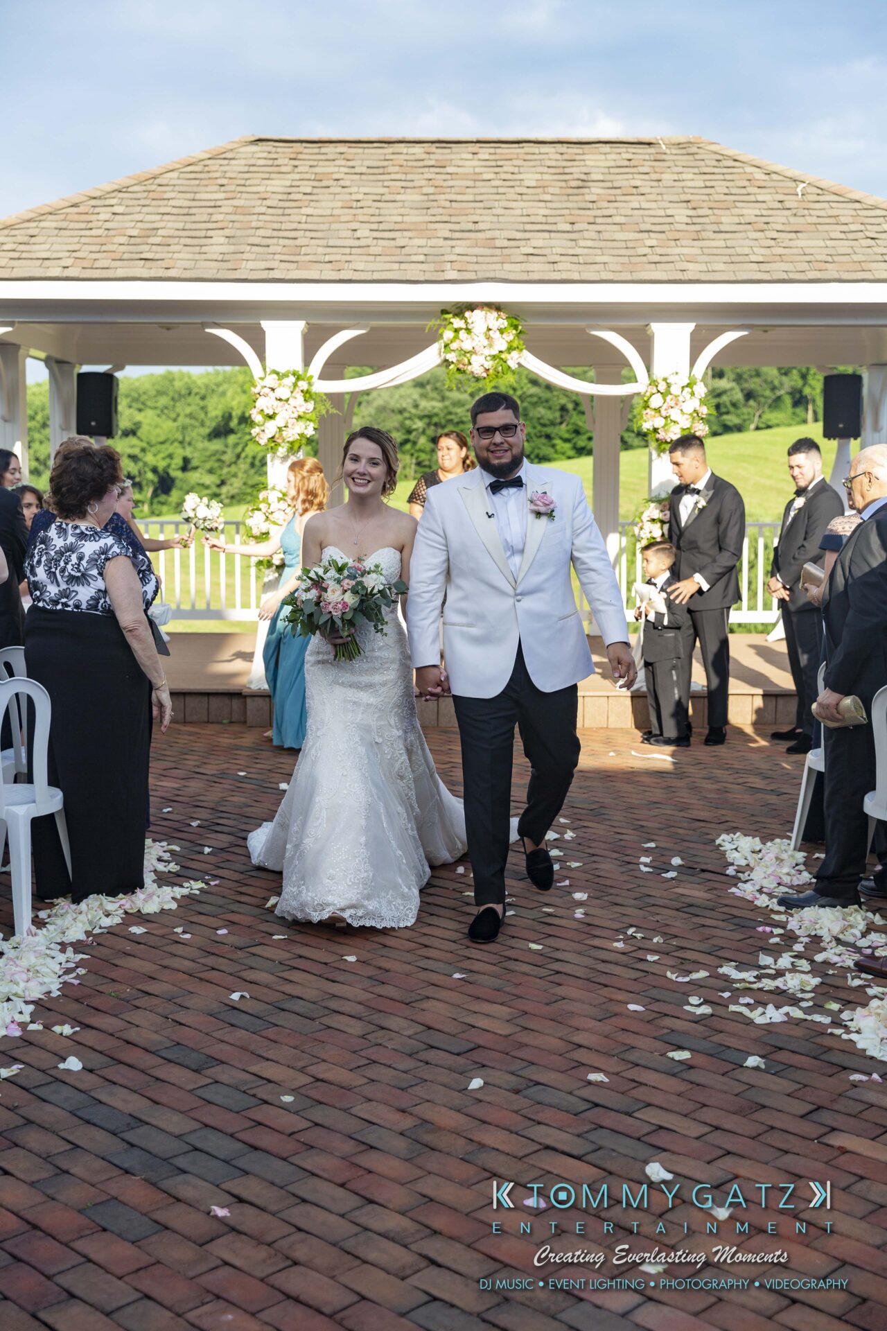 Kari-Jose-062318-CEREMONY-Low-Res-WM-331 - TOMMY GATZ ENTERTAINMENT A bride and groom walk down an outdoor aisle covered in rose petals, smiling at guests. Guests are seated on either side, and there is a gazebo in the background.