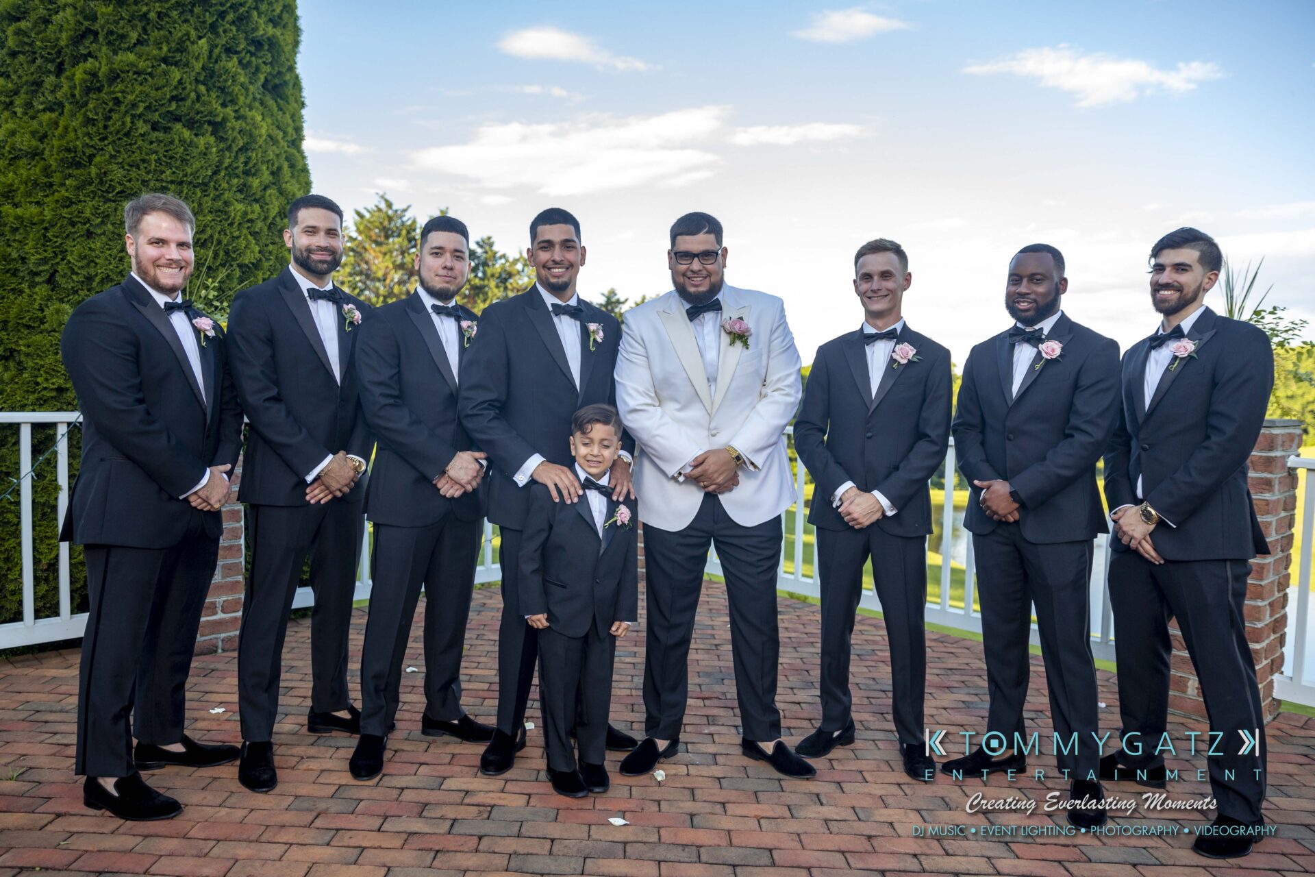 Kari-Jose-062318-FORMALS-Low-Res-WM-363 - TOMMY GATZ ENTERTAINMENT A group of men and a child, dressed in suits, pose outdoors on a brick patio. One man wears a white jacket, while the others wear black. Trees and a railing are visible in the background.
