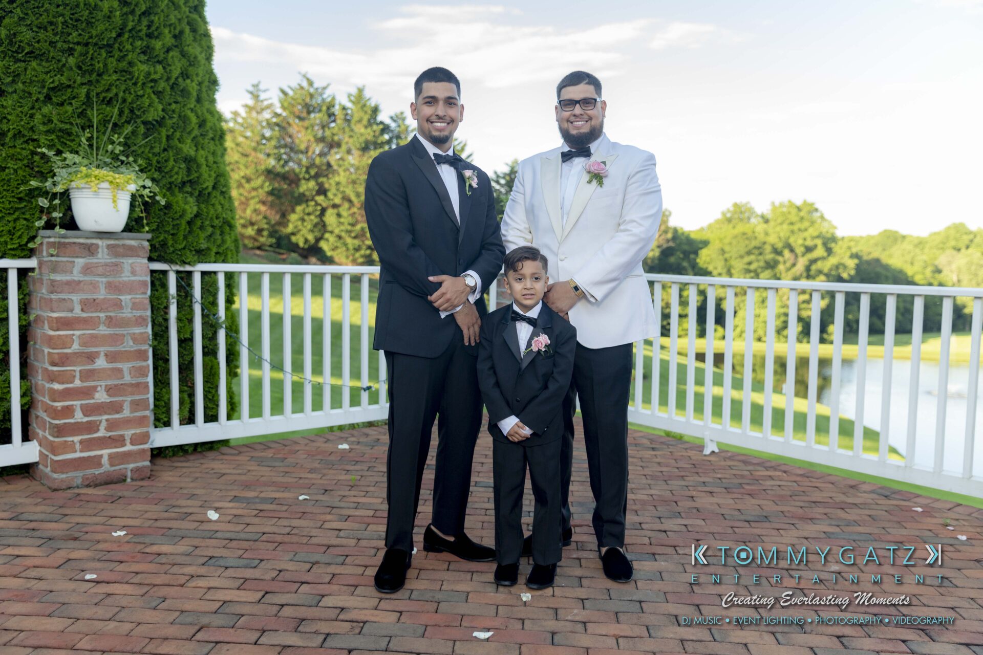 Kari-Jose-062318-FORMALS-Low-Res-WM-366 - TOMMY GATZ ENTERTAINMENT Three people in formal attire stand on a brick patio with a white fence and greenery in the background. Two adults are flanking a child in matching suits.