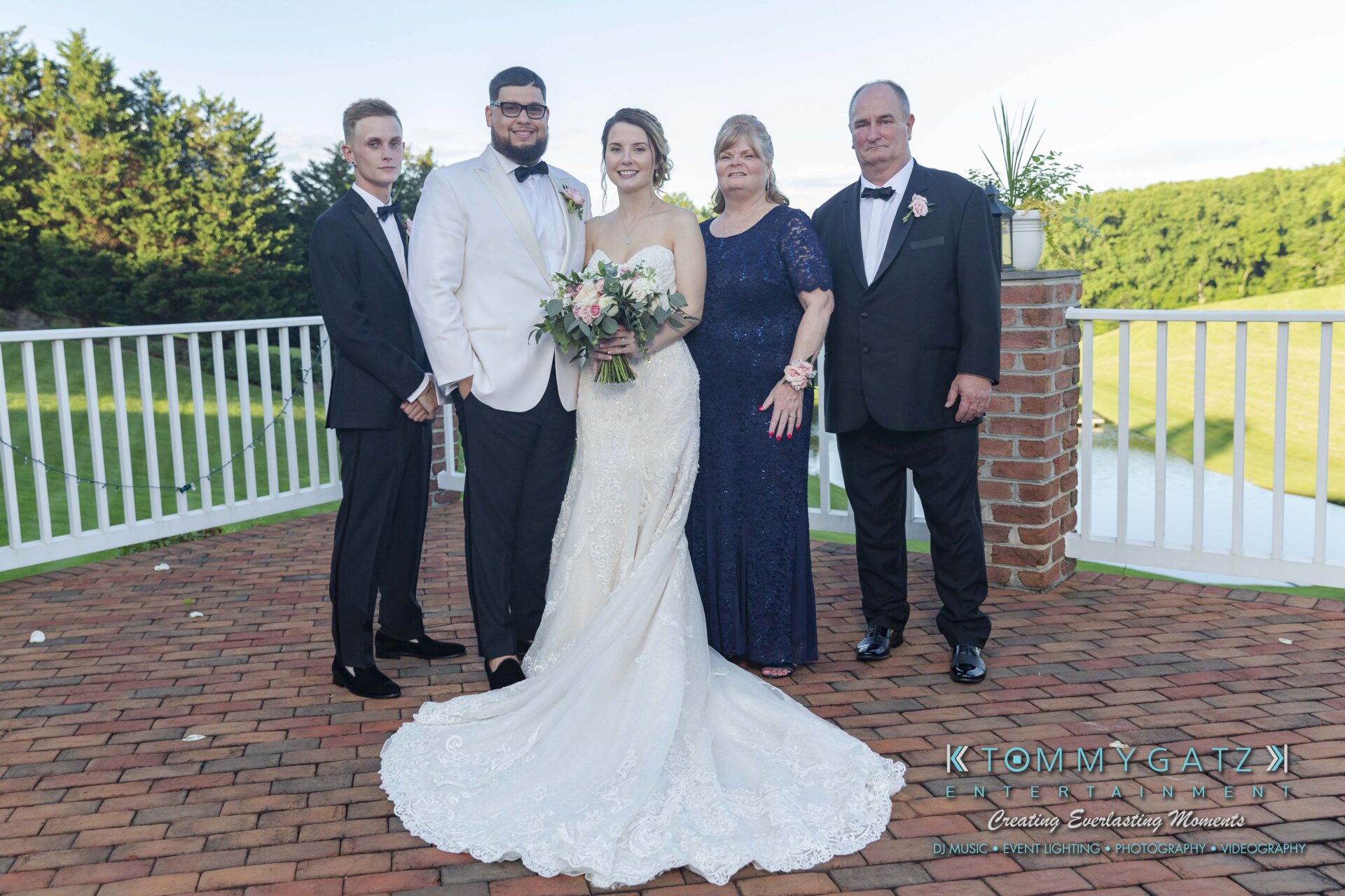 Kari-Jose-062318-FORMALS-Low-Res-WM-390 - TOMMY GATZ ENTERTAINMENT A wedding couple poses with three adults outdoors on a brick patio. The bride holds a bouquet, and everyone is dressed in formal attire. A green landscape is in the background.