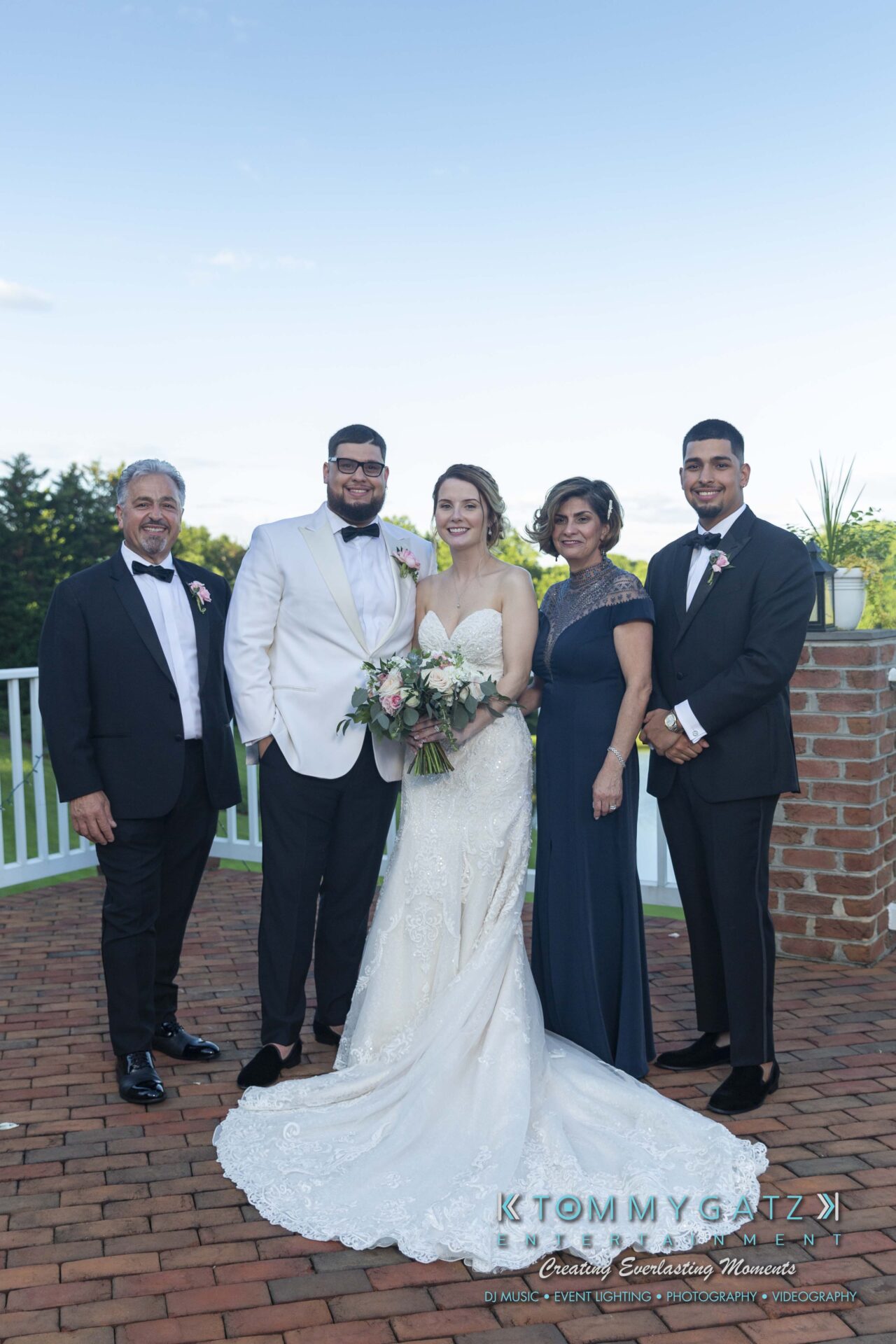 Kari-Jose-062318-FORMALS-Low-Res-WM-392 - TOMMY GATZ ENTERTAINMENT A bride and groom stand together with three other people on a brick patio, all dressed in formal attire.