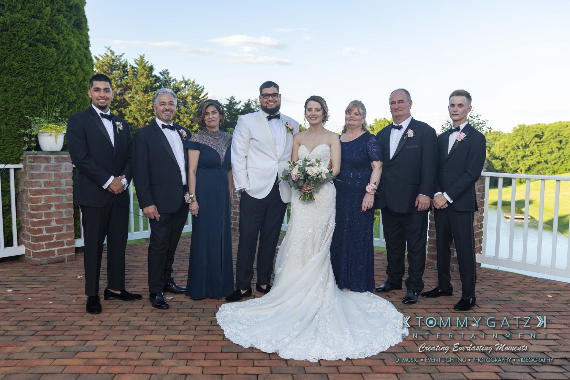 Kari-Jose-062318-FORMALS-Low-Res-WM-395 - TOMMY GATZ ENTERTAINMENT A bridal couple stands with six formally dressed individuals on a brick patio with a grassy backdrop.
