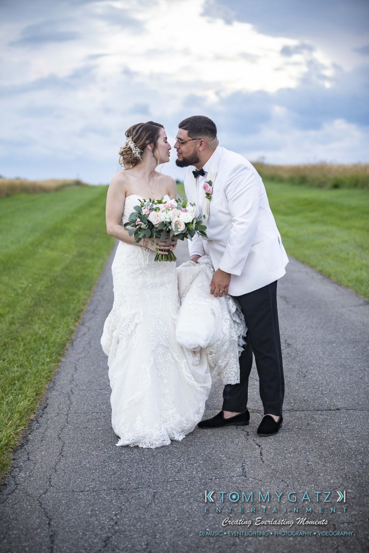 Kari-Jose-062318-FORMALS-Low-Res-WM-399 - TOMMY GATZ ENTERTAINMENT A bride and groom in wedding attire stand closely on a country road, holding a bouquet and smiling at each other.