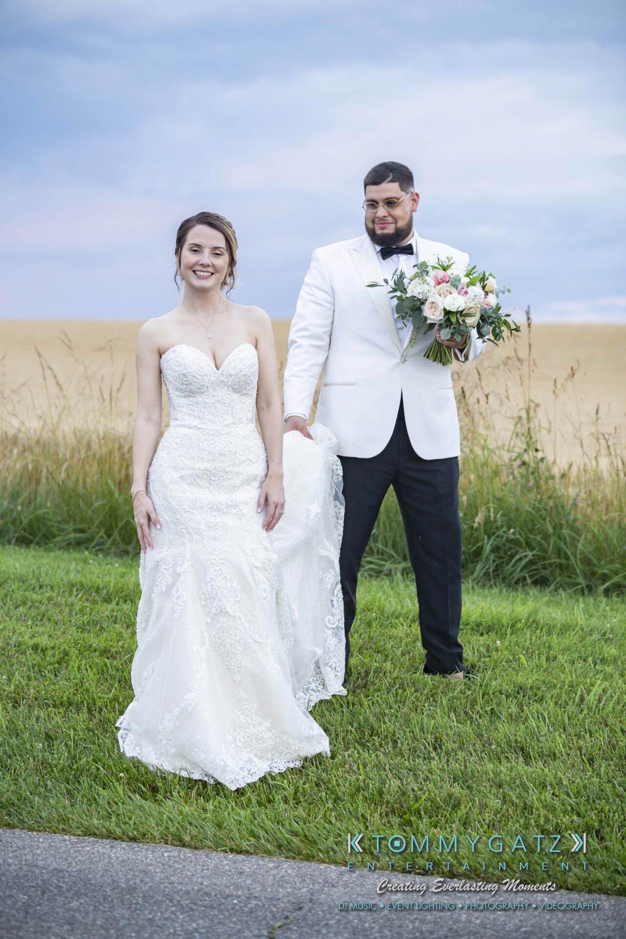 Kari-Jose-062318-FORMALS-Low-Res-WM-403 - TOMMY GATZ ENTERTAINMENT A bride and groom pose outdoors on grass, with the groom holding a bouquet. The sky is overcast, and a wheat field is in the background.