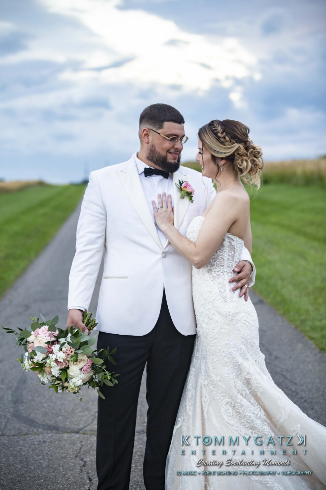 Kari-Jose-062318-FORMALS-Low-Res-WM-405 - TOMMY GATZ ENTERTAINMENT A bride and groom stand on a country road. The bride holds a bouquet of flowers, and they look at each other with smiles.