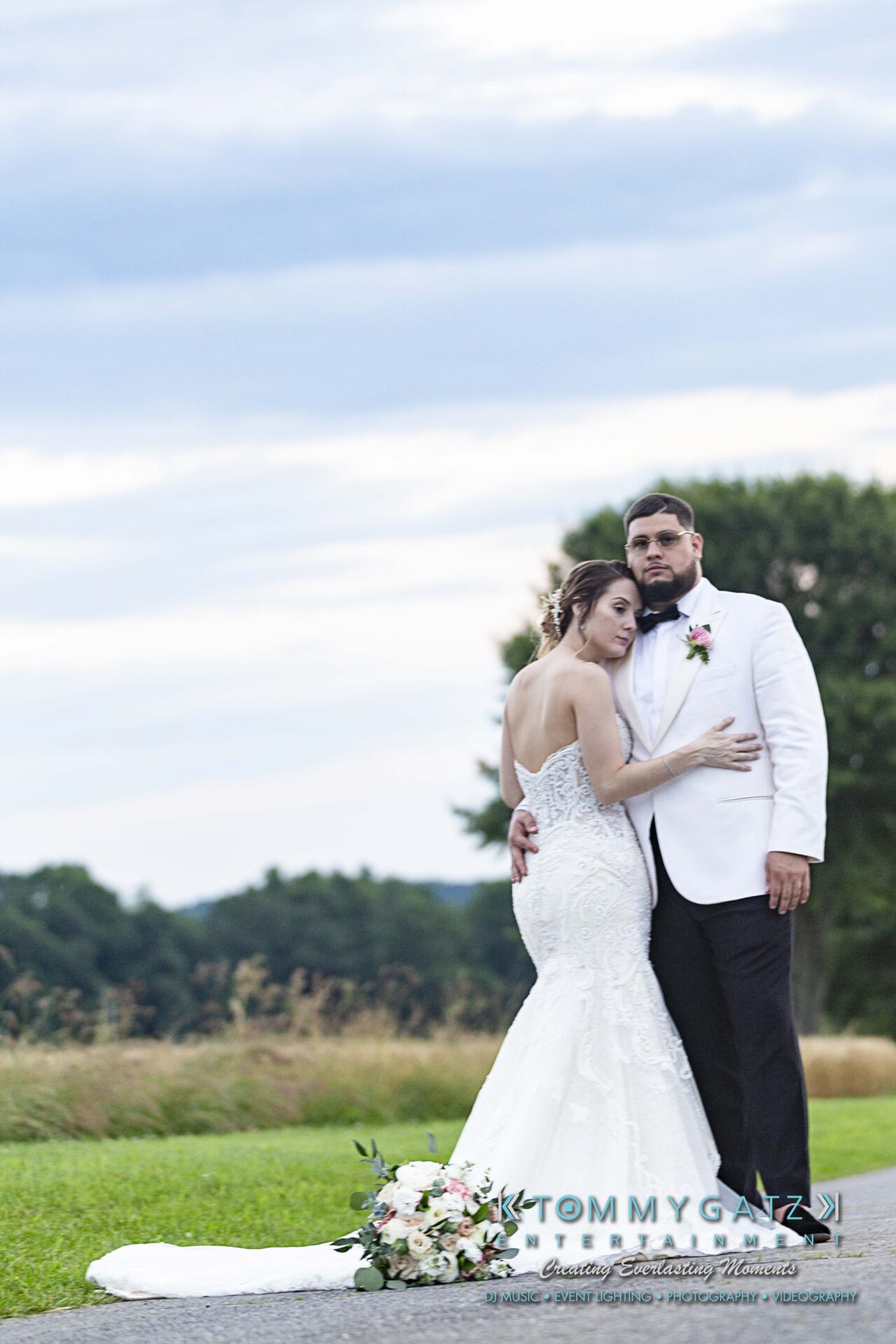 Kari-Jose-062318-FORMALS-Low-Res-WM-417 - TOMMY GATZ ENTERTAINMENT A couple in wedding attire stands on a grassy path, the bride in a white gown, the groom in a white jacket. A bouquet lies on the ground beside them.